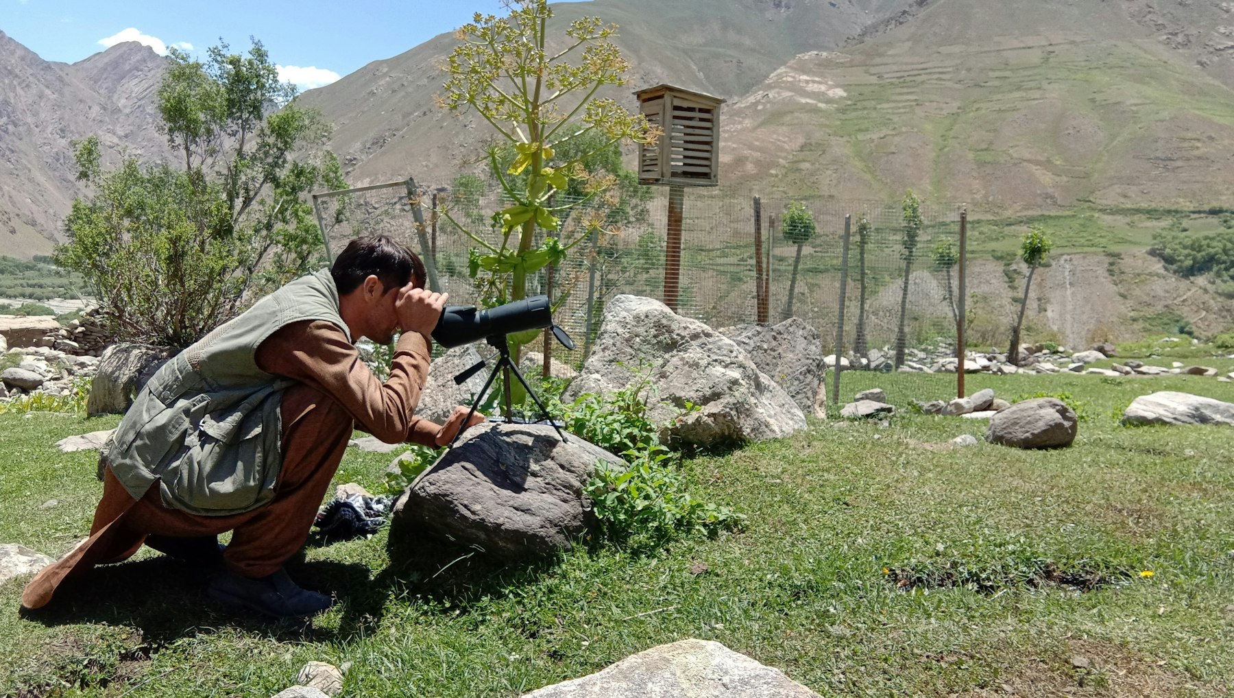 An AKAH weather monitoring volunteer in Arakht, Afghanistan kneels beside a weather monitoring post to look at a snow mast installed high on the mountain. Photo Credit: AKAH / Doug Chabot